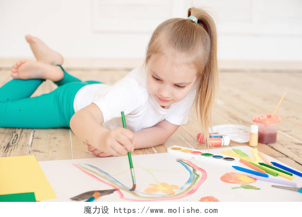 趴在地板上绘画的小女孩小金发女孩画在大白皮书, 而躺在地板上的室内